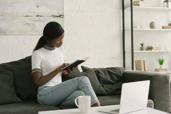 Jovem Africano americano freelancer leitura notebook enquanto sentado no sofá perto da mesa com laptop — Fotografia de Stock