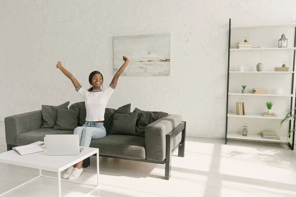 Feliz afroamericano freelancer estiramiento mientras está sentado en el sofá cerca de la mesa con el ordenador portátil en la amplia sala de estar - foto de stock