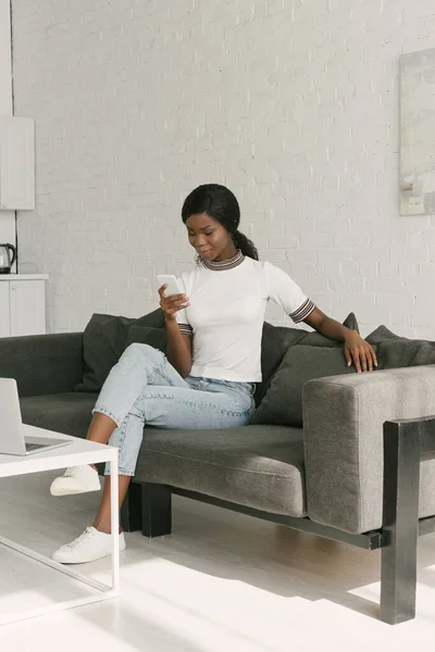 Молодой африканский американский фрилансер болтает на смартфоне, сидя за столом с ноутбуком на кухне — стоковое фото