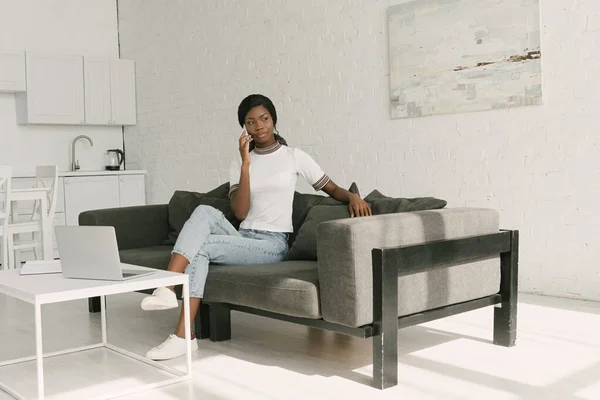Вдумчивый африканский фрилансер, говорящий на смартфоне, сидя на диване рядом со столом с ноутбуком — стоковое фото