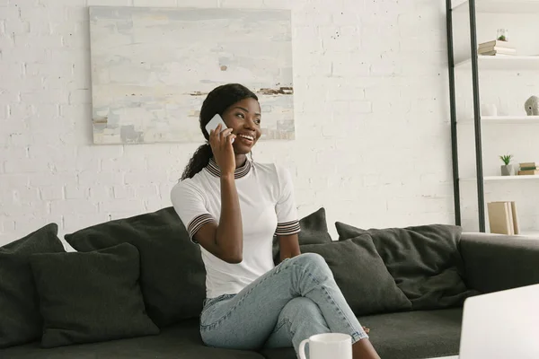 Menina americana africana alegre sentado no sofá, falando no smartphone e olhando para longe — Fotografia de Stock