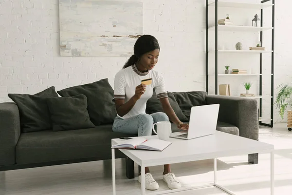 Chica afroamericana bonita usando el ordenador portátil y la celebración de la tarjeta de crédito mientras está sentado en el sofá en casa - foto de stock