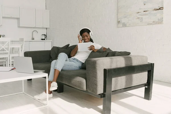 Heureux freelance afro-américain dans les écouteurs sans fil en utilisant une tablette numérique tout en étant assis sur le canapé près de la table avec ordinateur portable dans la cuisine — Photo de stock