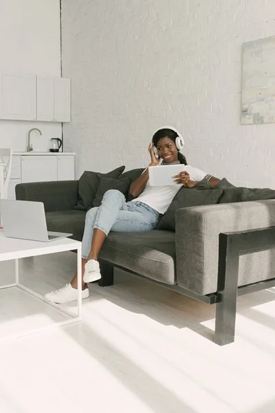 Веселий афроамериканський фрілансер у бездротових навушниках, що сидить на дивані та тримає цифровий планшет біля столу з ноутбуком — стокове фото