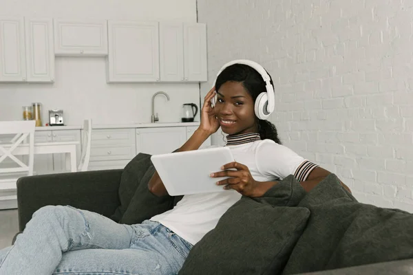 Menina americana africana feliz em fones de ouvido sem fio sentado no sofá na cozinha e segurando tablet digital — Fotografia de Stock