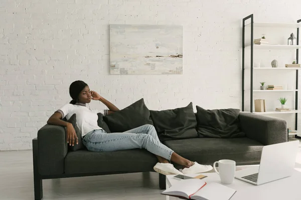 Pensativo afroamericano freelancer descansando en el sofá cerca de la mesa con portátil en la sala de estar - foto de stock