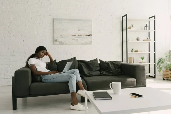 Thoughtful african american freelancer sitting on sofa with laptop in spacious living room — Stock Photo