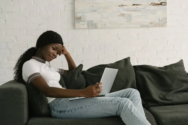 Sonriente afroamericano freelancer utilizando el ordenador portátil mientras está sentado en el sofá en casa - foto de stock