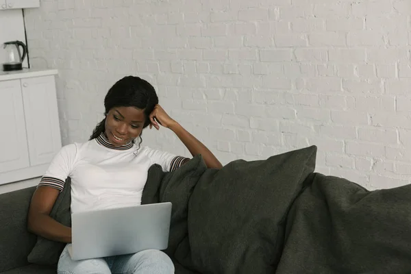 Sonriente afroamericano freelancer sentado en el sofá y utilizando el ordenador portátil - foto de stock