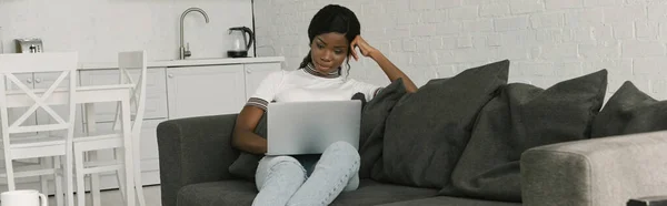 Horizontales Bild einer aufmerksamen afrikanisch-amerikanischen Freelancerin, die am Laptop auf dem Sofa in der Küche arbeitet — Stockfoto