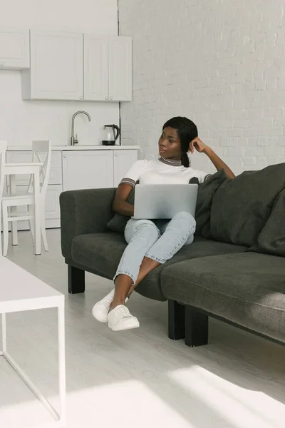 Pensive african american freelancer sitting on sofa with laptop and looking away — Stock Photo