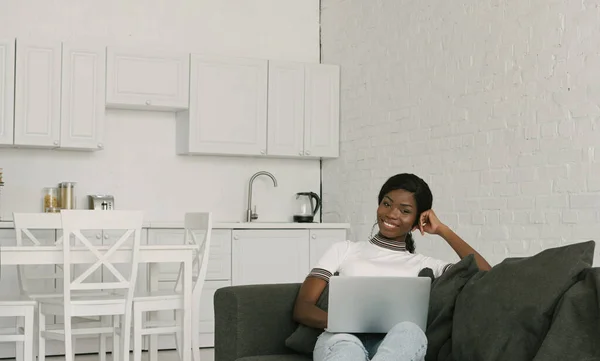 Happy african american freelancer sitting on sofa with laptop and smiling at camera — Stock Photo