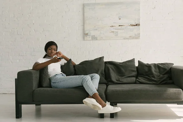 Приваблива афроамериканська дівчина тримає телевізійний пульт дистанційного керування, сидячи на дивані вдома — стокове фото