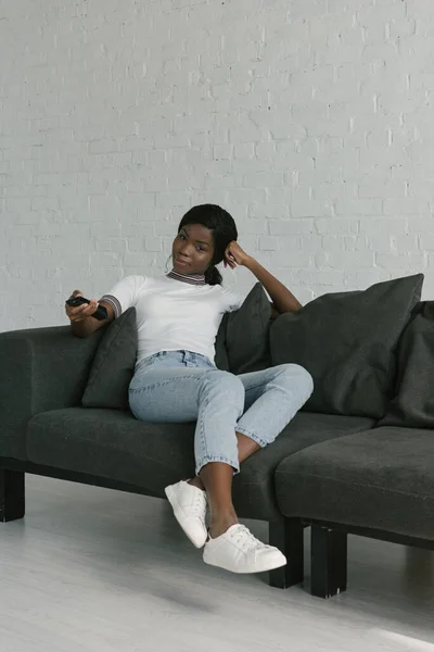 Довольно африканская американская девушка держит телевизор пульт дистанционного управления, сидя на диване дома — стоковое фото