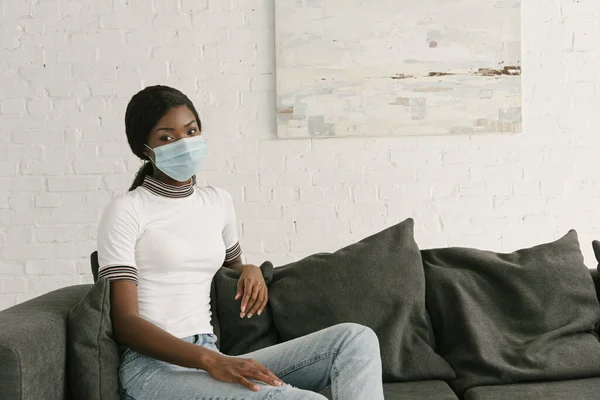 Chica afroamericana pensativa en máscara médica sentado en sofá gris en casa - foto de stock