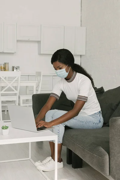 Молодой африканский американский фрилансер в медицинской маске, работающий на ноутбуке на кухне — стоковое фото