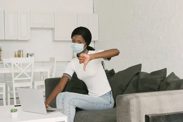 Young african american freelancer in medical mask showing thumb down while sitting near laptop in kitchen — Stock Photo