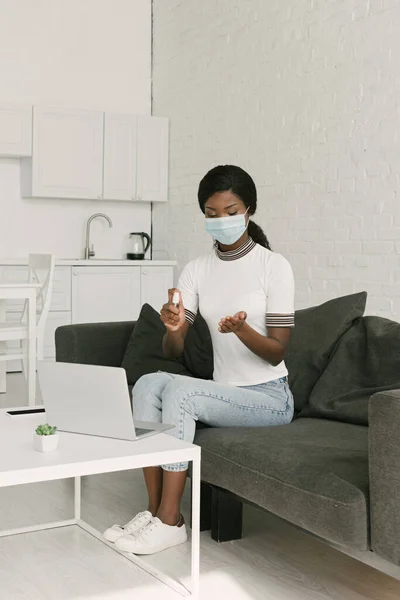 Giovane freelance afroamericano in maschera medica spruzzando antisettico sulle mani mentre seduto vicino al computer portatile in cucina — Foto stock