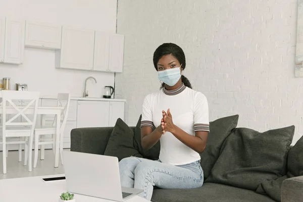 Joven afroamericano freelancer en máscara médica desinfectando manos mientras está sentado cerca de la computadora portátil en la cocina - foto de stock