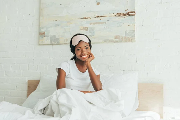 Alegre afroamericana chica con máscara de sueño en la frente sentado en la cama y sonriendo a la cámara - foto de stock