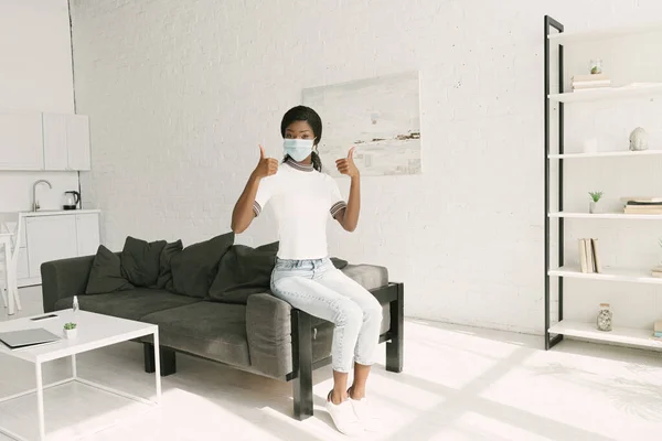 Молода афроамериканська дівчина в медичній масці показує великі пальці вгору, сидячи на сірому дивані на кухні — стокове фото