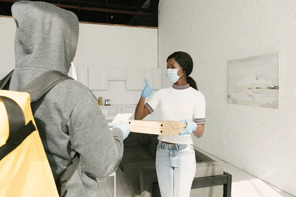 Afro-américaine fille en masque médical et gants en latex prendre des boîtes avec pizza de livreur — Photo de stock