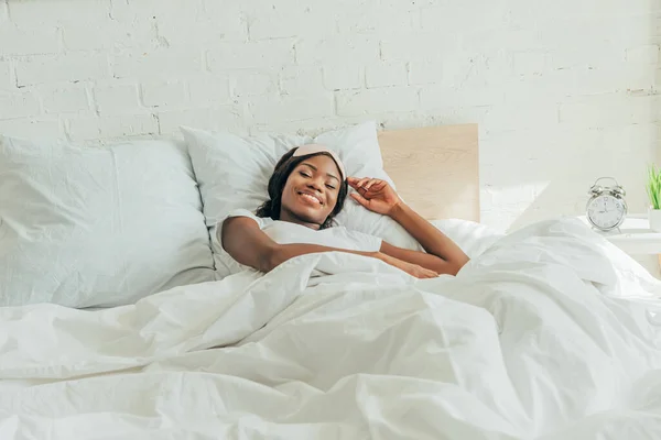 Feliz afroamericana chica con máscara de sueño en la frente acostado en la cama y sonriendo a la cámara - foto de stock