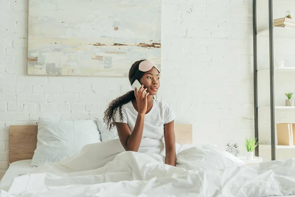 Reflexiva chica afroamericana con máscara de sueño en la frente hablando en el teléfono inteligente mientras está sentado en la cama - foto de stock