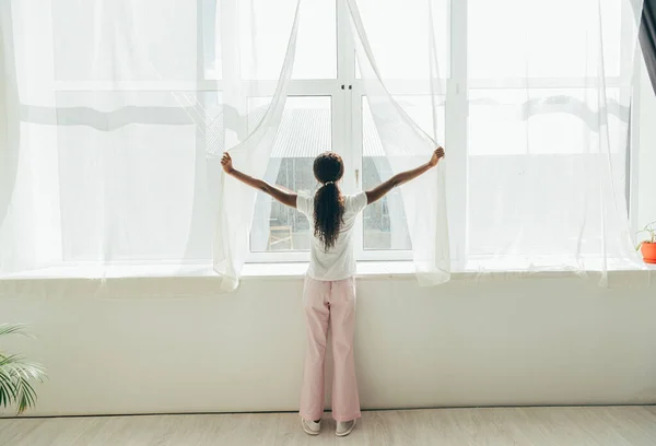 Vue arrière de la fille afro-américaine en pyjama ouvrant rideaux de fenêtre dans la lumière du soleil — Photo de stock