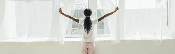 Vista trasera de chica afroamericana en pijama abriendo cortinas de ventana bajo el sol, imagen horizontal - foto de stock