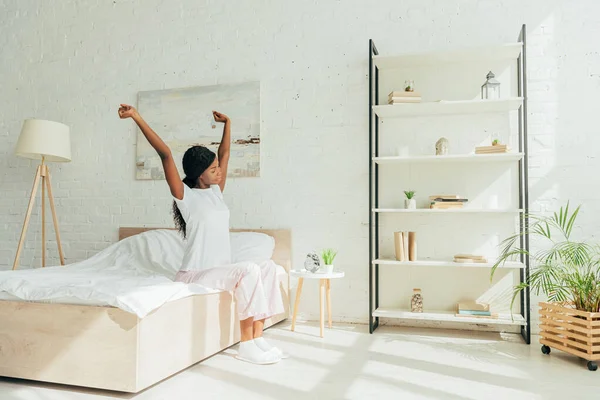 Afroamericana chica en pijama sentado en amplio dormitorio y estiramiento con las manos levantadas - foto de stock