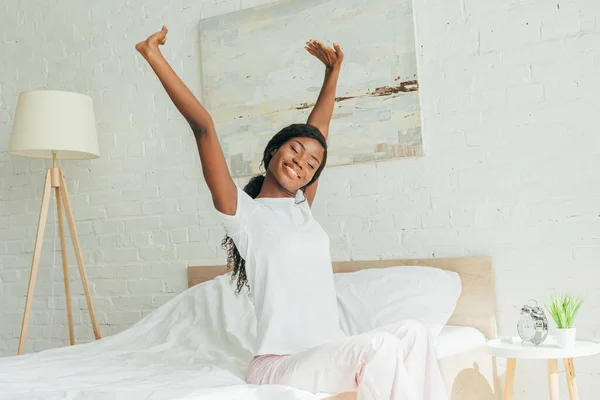 Усмішка африканської американки в піжамі сидячи на ліжку з закритими очима і розтягуючись з піднятими руками. — стокове фото