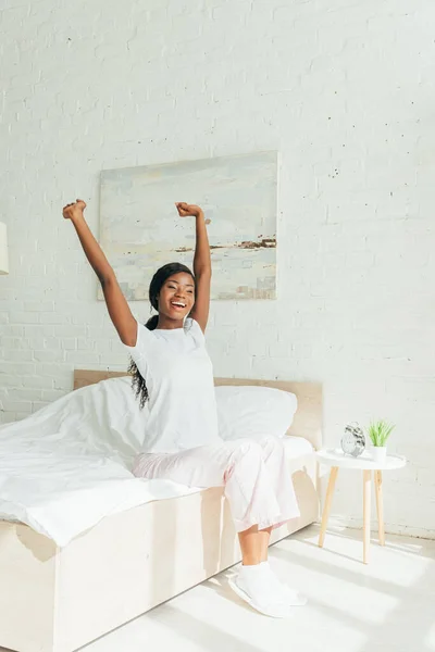 Menina americana africana feliz em pijama alongando-se com as mãos levantadas enquanto sentado na cama — Fotografia de Stock