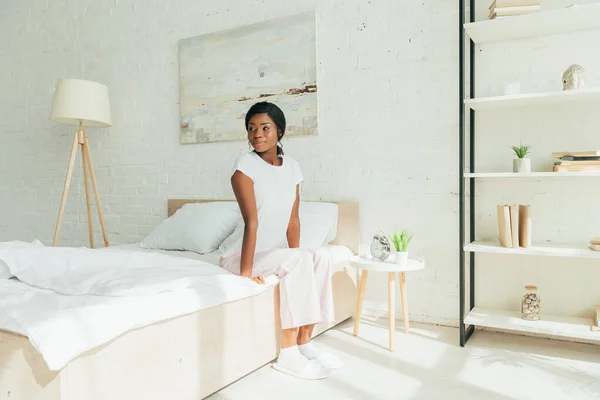 Bela menina americana africana em pijama sentado na cama e olhando para longe de manhã — Fotografia de Stock