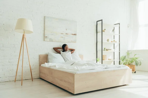Feliz afroamericana chica sentada en la cama con las manos detrás de la cabeza en amplio dormitorio - foto de stock