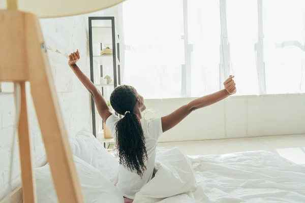 Vue de dos de la fille afro-américaine étirant avec les mains levées tout en étant assis dans le lit — Photo de stock