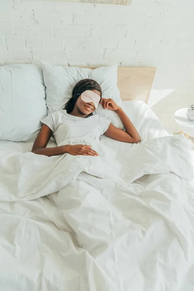 Vista ad alto angolo di sorridente ragazza africana americana sdraiata a letto in maschera del sonno — Foto stock