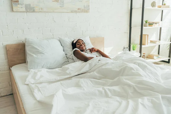 Menina americana africana feliz com máscara de sono na testa sorrindo enquanto deitado na cama — Fotografia de Stock