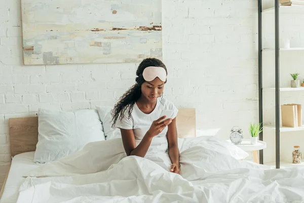 Jolie africaine américaine fille avec masque de sommeil sur le front bavarder sur smarthone tout en étant assis dans le lit — Photo de stock