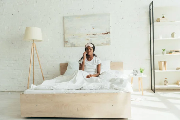 Mujer afroamericana joven con máscara de sueño en la frente sentada en la cama mientras habla en el teléfono inteligente - foto de stock