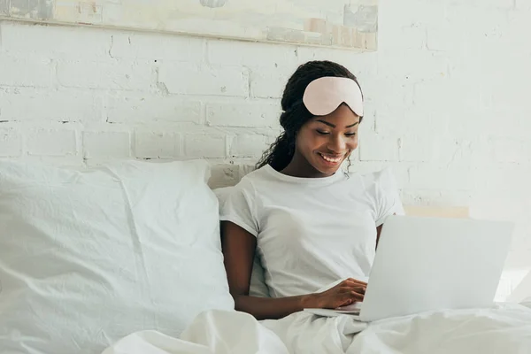 Freelancer afro-americano sorridente com máscara de sono na testa usando laptop na cama — Fotografia de Stock