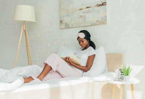 Atenciosa menina afro-americana com máscara de sono na testa leitura livro enquanto sentado na cama — Fotografia de Stock