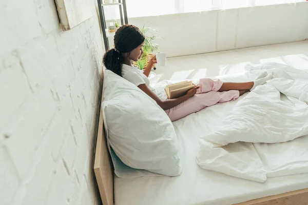 Vista de ángulo alto de la chica afroamericana sentada en la cama y leyendo libro - foto de stock