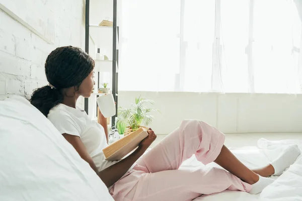African american girl in pajamas holding book and looking away while sitting in bed — Stock Photo