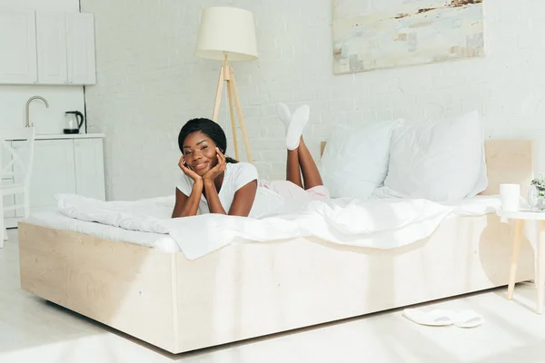 Menina americana africana feliz sorrindo para a câmera enquanto deitado em roupa de cama branca — Fotografia de Stock