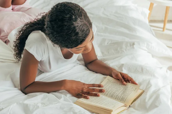 Vista aérea de la joven afroamericana leyendo libro en la cama - foto de stock