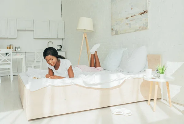 Linda menina americana africana deitada na cama branca e livro de leitura — Fotografia de Stock