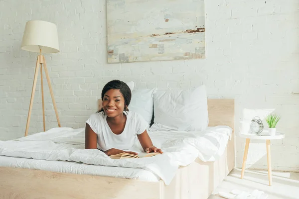 Fröhliches afrikanisch-amerikanisches Mädchen liegt mit Buch im Bett und lächelt in die Kamera — Stockfoto