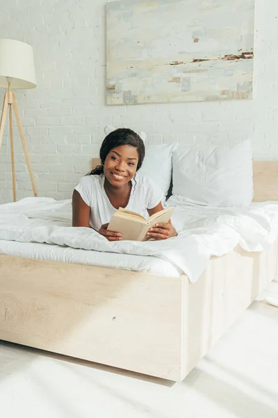Felice ragazza africana americana sdraiata con libro aperto su biancheria da letto bianca — Foto stock