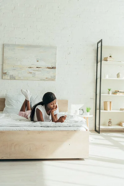 Africano americano ragazza sdraiata a letto e chattare su smartphone in spaziosa camera da letto — Foto stock